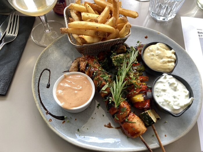 Grillspett med grönsaker och såser på en tallrik, serverat med pommes frites och ett glas vin.