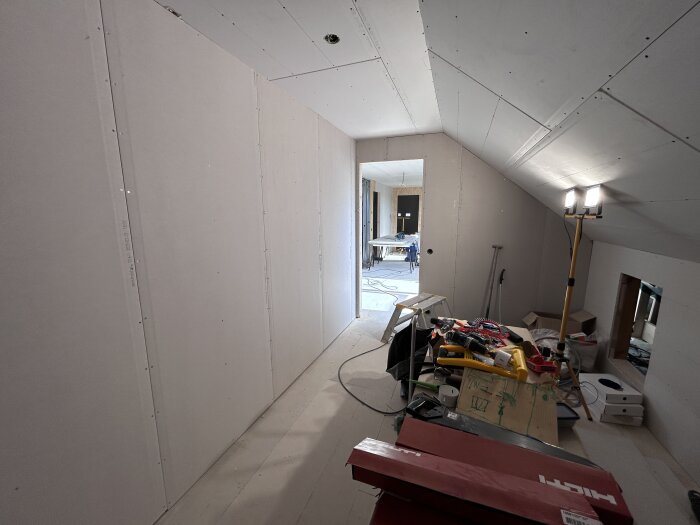 Byggarbetsplats med ett halvfärdigt rum; gipsskivor monterade på väggar och tak; byggmaterial och verktyg på golvet och en arbetslampa lyser upp rummet.