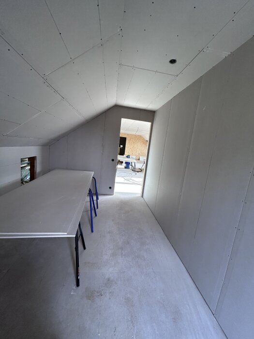 Rum under konstruktion med gipsskivor på väggar och tak, bord stödt på bockar; öppning mot angränsande rum syns i bakgrunden.