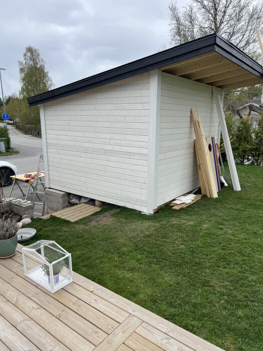 Vitmålad redskapsbod med svart tak i trädgård; byggmaterial och träpaneler står lutade mot bodens vägg.
