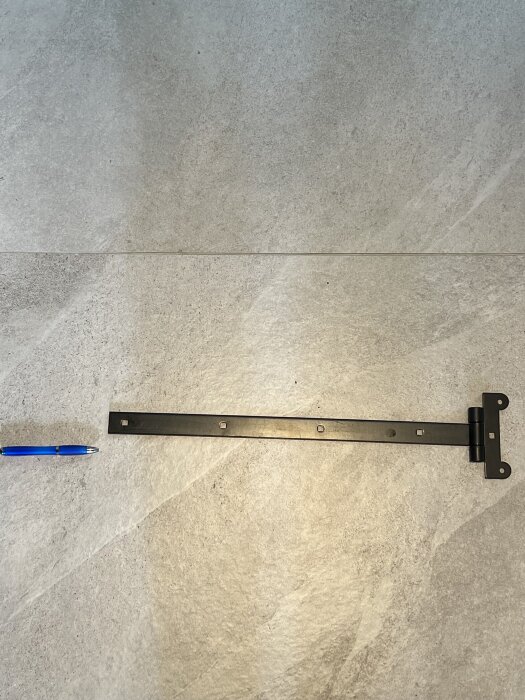 Svart hakgångjärn ligger på ett stengolv bredvid en blå kulspetspenna för storleksjämförelse.