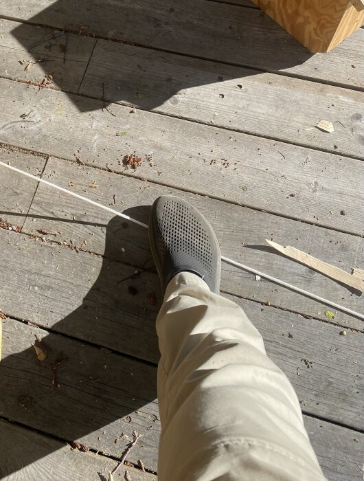 Fot på brygga pressar linan mot marken för att stabilisera ämne vid tillverkning av en långbåge; improviserat arbetsmoment vid täljning.