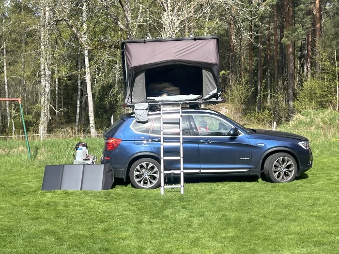 Blå bil med tält på taket, uppfällt med stege bredvid. Vikbar solpanel, gelbatteri och kompressorkyl syns på marken.