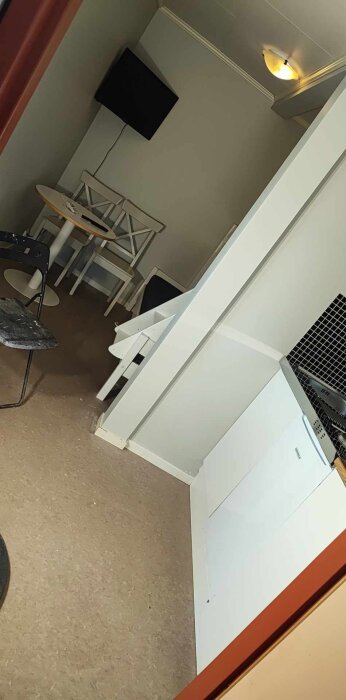 Minimalistiskt kök nästan klart, med vita skåp, bord, stolar och en svart väggmonterad tv, redo för sista steg som kökskran och hyllor.