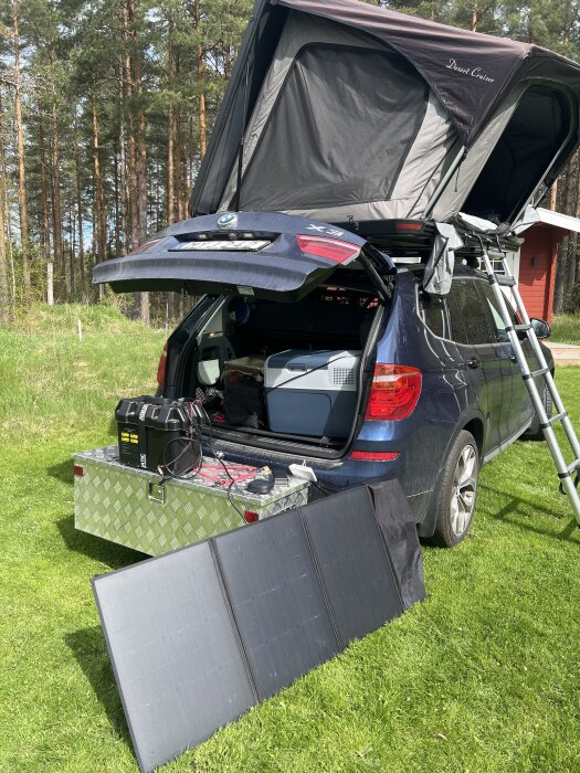 Bil med bakluckan öppen, solpaneler och en uppfälld taktält i skogsmiljö.