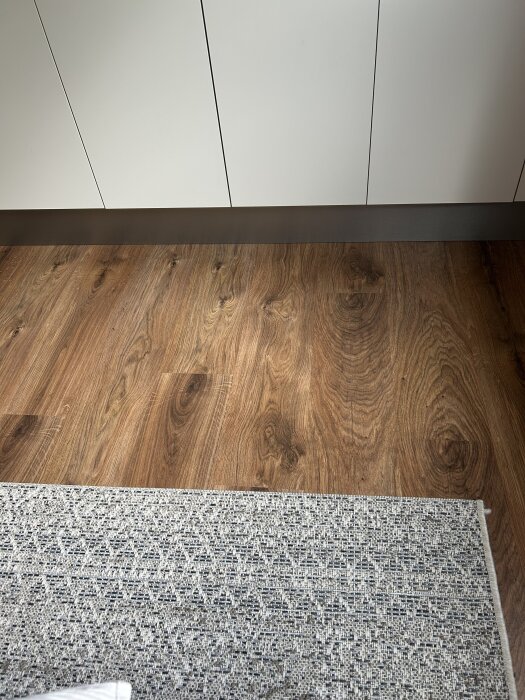 Mörkbrunt Ikea Tundra laminatgolv med träådring, delvis täckt av en gråvit matta framför vita skåp.
