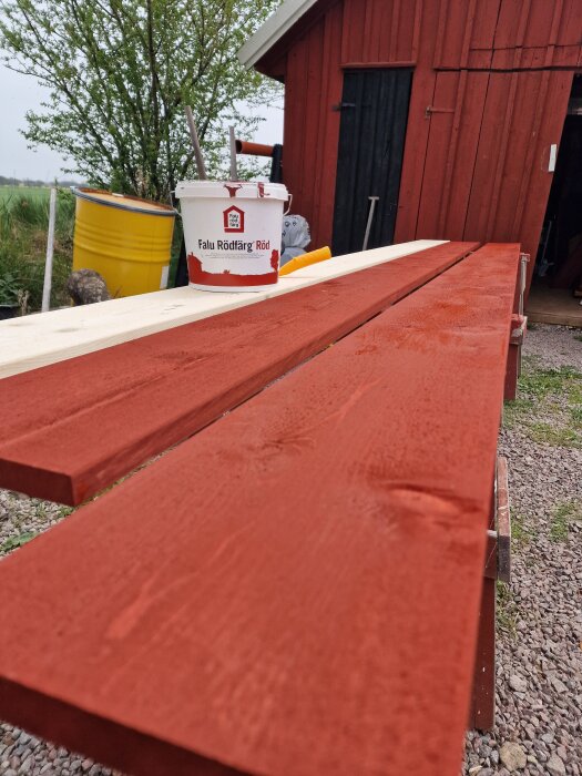 Rödbrunmålad träpanel står på mark med en burk Falu Rödfärg, omslutet av grönska och en röd träbyggnad i bakgrunden.