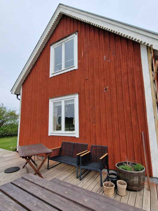 Röd träfasad med två fönster, trätrall, cafébord, bänk och krukväxter på altanen. Renovering och tilläggsisolering pågår.