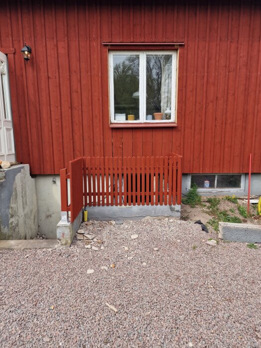 Röd träfasad med ett vitt fönster, grusad yta framför och ett nytt rött staket kring en nedgång mot källaren. Gavelprojekt med återbrukat material.