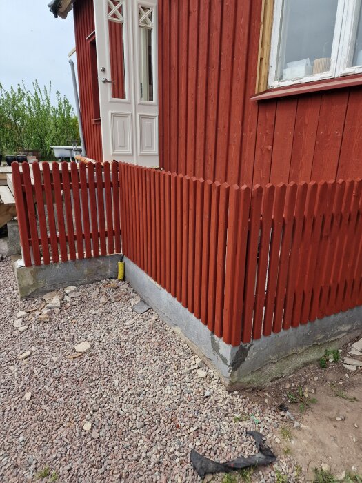 Röd träfasad med ny brädfodring och ett staket, renovering pågår. Nijstenraveler en entré med vita ytterdörrar och gruslagd gång bredvid huset.