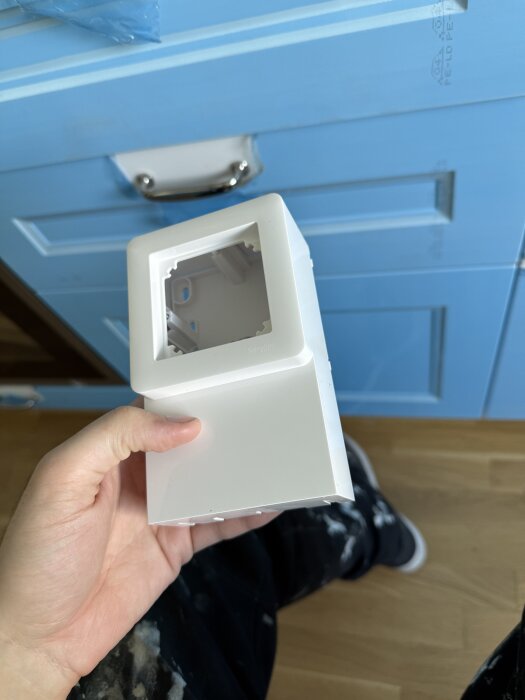 Vit tom sockeldosa, Exxact, hålls i handen framför en ljusblå skåplucka. Dosan saknar lock och placeras ovanpå golvet.