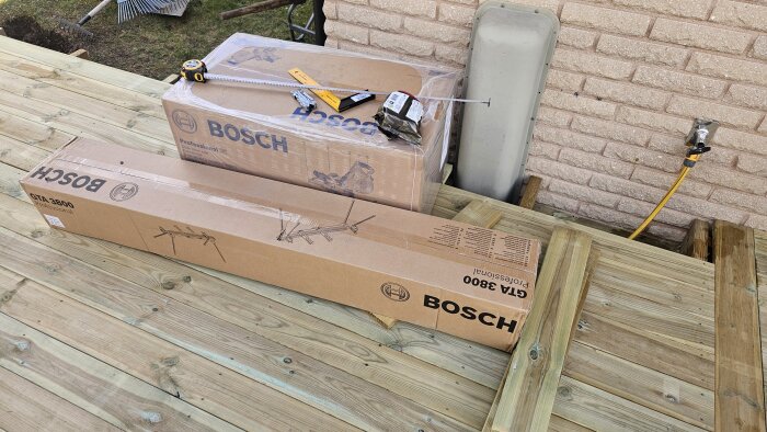 Förpackningar med Bosch kap- och gersåg GCM8SJL och stativ GTA 3800 på trätrall med måttband och verktyg ovanpå.