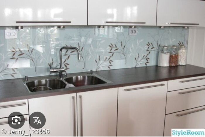 Köksbänk med rostfri diskho, vitrinluckor och glaspanel med bladmönster ovanför diskbänken.
