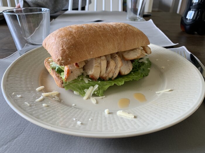 Sandwich med grillad kyckling och sallad på en tallrik, omgiven av riven ost och några droppar dressing.