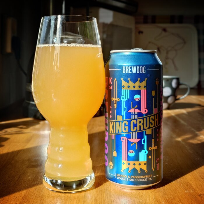 Ett glas med en gul dryck bredvid en färgglad burk med texten "King Crush" på ett bord.