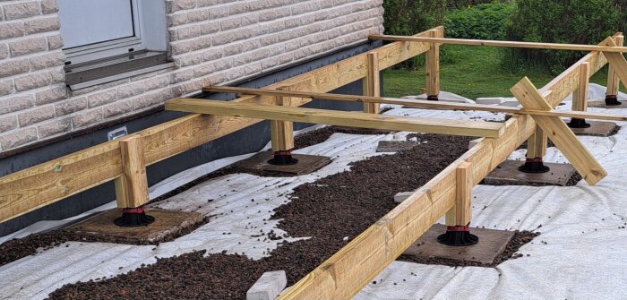 Trästomme för altanbygge med bärlinor på terassfotsben, markduk och singel synligt under plattor intill husvägg.