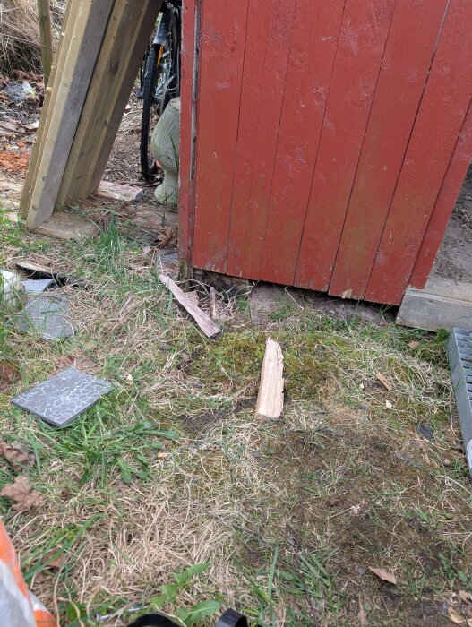 Röd vedbod med trasig grund och jordnära brädor, omgiven av gräs och träbitar. En cykel skymtas i bakgrunden.