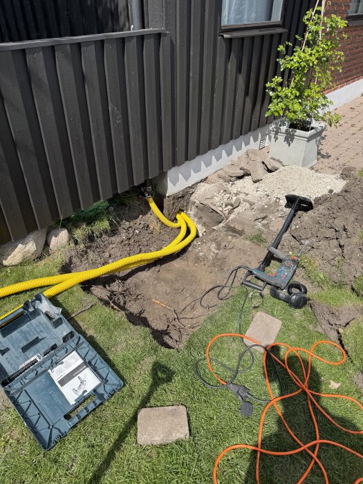 Gul slang installerad i marken nära husvägg, omgiven av verktyg och elsladdar. Förberedelser för framtida belysning och laddboxar.