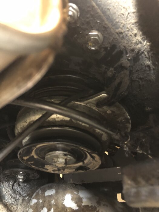 Närbild på åkgräsklippares motorområde där drivremmar och remhjul syns, med smutsiga delar runt om.