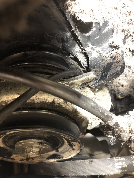 Åkgräsklipparens drivrem och remhjul med omgivande damm och kabel i närbild under motorutrymmet.