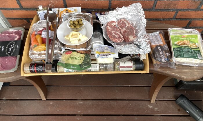 Matvaror för grillning, inklusive kött, ostar, paprikor, potatisgratäng, kryddor och grillkorv, upplagda på en bricka utomhus.