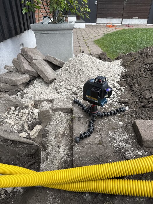 Gul slang i jord vid byggarbetsplats; laser på stativ och uppgrävt material syns.