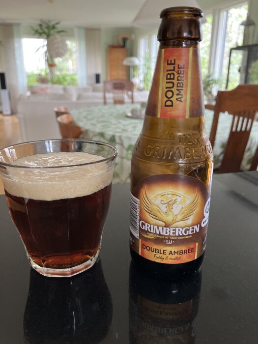 Grimbergen Double Ambree ölflaska och glas med skummande mörkbrun öl på ett svart bord i ett ljust vardagsrum.