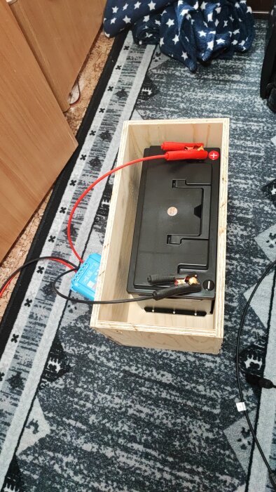Låda av trä med bilbatteri och anslutna kablar placerad på golvmatta, med batterimonitor synlig bredvid.