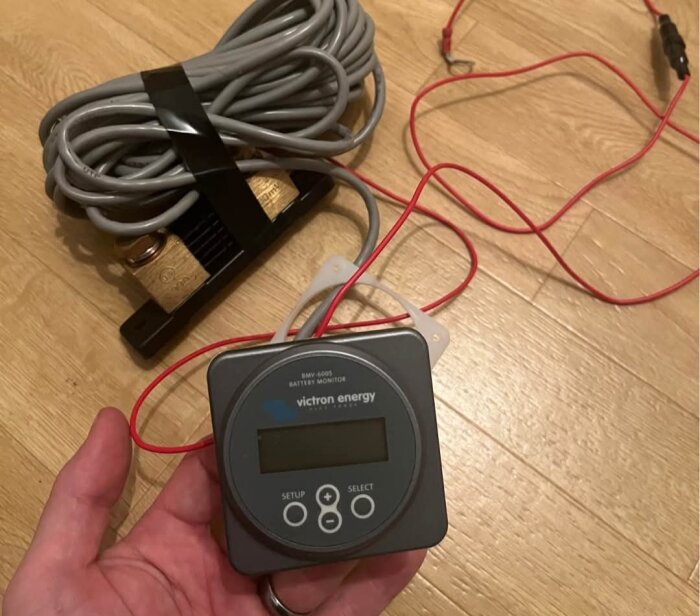 Hand håller Victron batterimonitor med röd kabel, shunt och grå kabel syns på golvet.