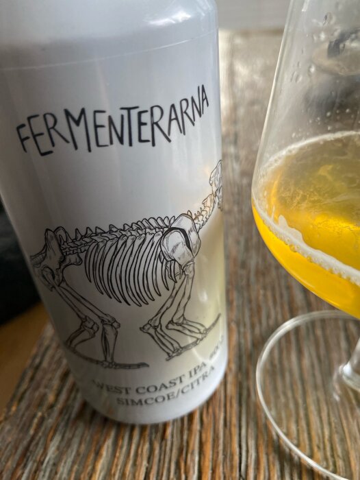 Burk med Fermenterarna West Coast IPA, illustrerad med ett skelett, bredvid ett halvt fullt glas gyllene öl på ett träbord.