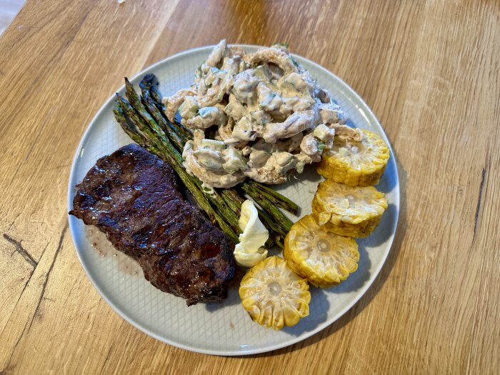 Tallrik med grillad biff, majs, sparris och potatissallad med gurka, rödlök och dill på en träbordsyta.