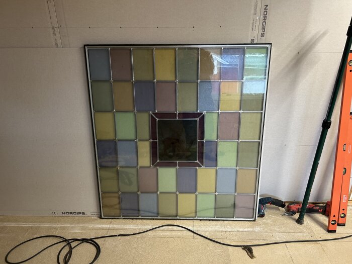 Blyinfattad glasskiva med färgglada rutor, centralt motiv oklart, omgiven av verktyg och byggmaterial i bakgrunden.