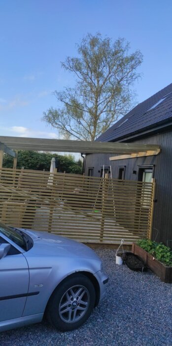 Bilden visar en vägghängd pergola byggd på en altan, med två stödjande stolpar och en träpanel. En bil och grusgård syns i förgrunden.