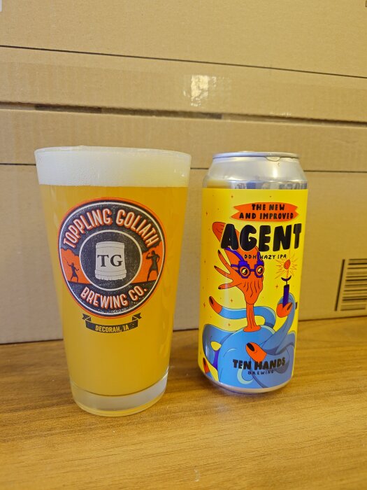 Ölglas med Toppling Goliath-logga bredvid en burk Ten Hands Agent DDH Hazy IPA, placerade på en träyta med kartongbakgrund.