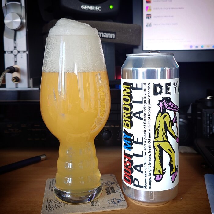 Ett glas med skummande ljus öl bredvid en burk med etiketten "Dust My Broom Pale Ale" från Deya, på ett skrivbord med teknisk utrustning.