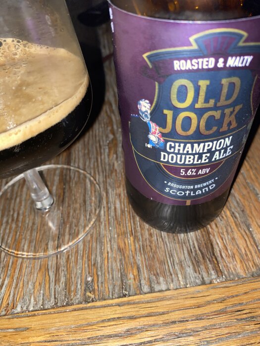 Flaska med etikett för Old Jock Champion Double Ale och ett glas mörk öl på ett träbord.