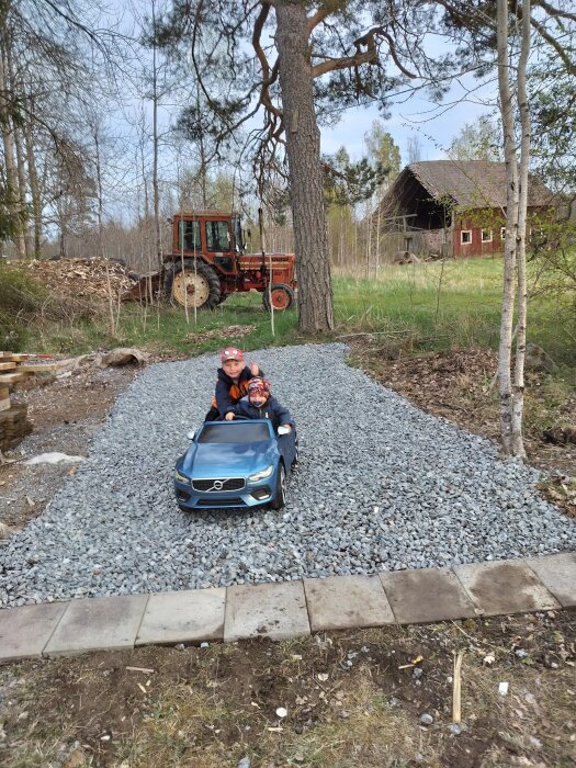 Två barn i en leksaksbil på en grusuppfart framför en röd traktor och en lada i skogsmiljö.