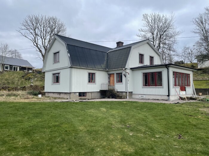 En vit tvåvåningsvilla med svart tak och röda fönsterkarmar, omgiven av gräsmatta och träd, under molnig himmel.