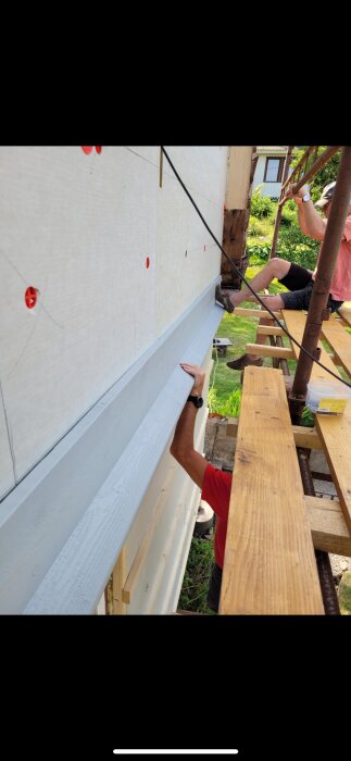 Person arbetar på ställning och monterar fasadplanka på husvägg med snedsågad droppnäsa, skruvad för stabilitet.