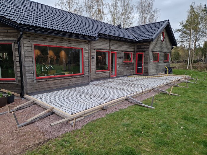 Förberedd gjutform för altan vid träfasadhus med rödbruna detaljer, el och tomrör dragna, omgiven av gräsmatta och träd.