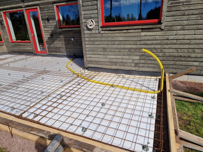 Förberedd yta för gjutning av altan med armeringsjärn och gulfärgat rör för kommande el- och vatteninstallation, intill träfasad med röda fönster och dörrar.