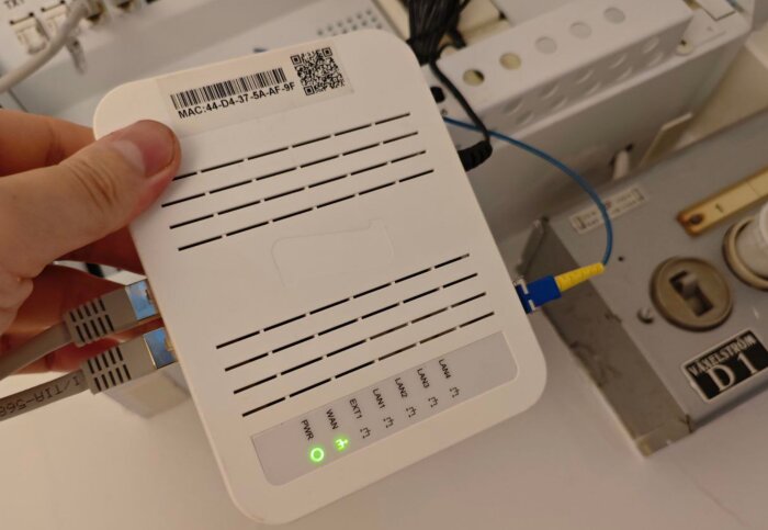 En hand håller en vit router med flera kablar anslutna, en grön strömindikator lyser.