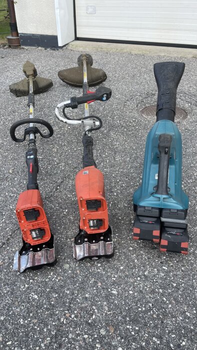 Tre trimmers med batteridrivna motorer placerade på en asfalterad gårdsplan. Två har orangeröda motorer och en har en blå motor.