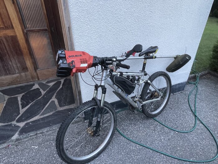 Cykel med en motor och en trimmer monterad på styret, parkerad mot en vit vägg bredvid en dörr.