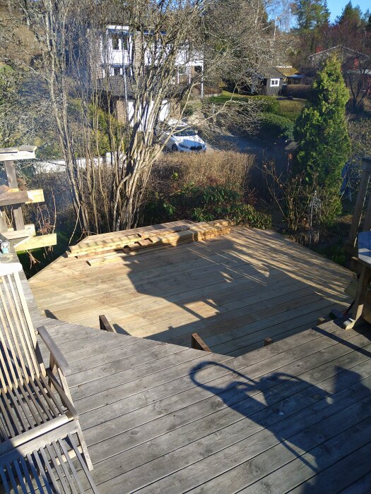 Trädäck under konstruktion med omkringliggande trädgård och hus i bakgrunden. Solbelyst trästruktur och sittplats i förgrunden.