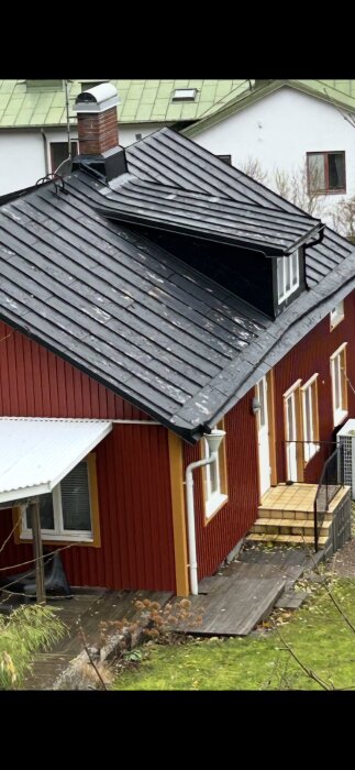 Svartmålat plåttak på ett rött hus med färgskiftningar och vit skorsten, omgärdat av en grön trädgård.