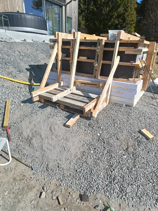 Byggkonstruktion med träbalkar och stödfästen på grusgrund nära ett hus med stora glasfönster och badtunna i bakgrunden.