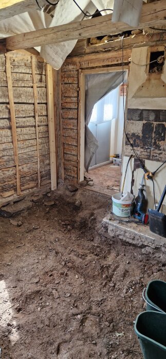 Renovering av gammalt hus med blottade timmerväggar, jordgolv och byggmaterial; synliga verktyg och utrustning under arbete.