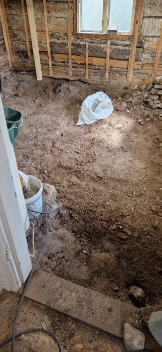 Renoveringsprojekt med uppgrävt jordgolv i ett timmerhus, synliga väggbalkar och byggmaterial på golvet.