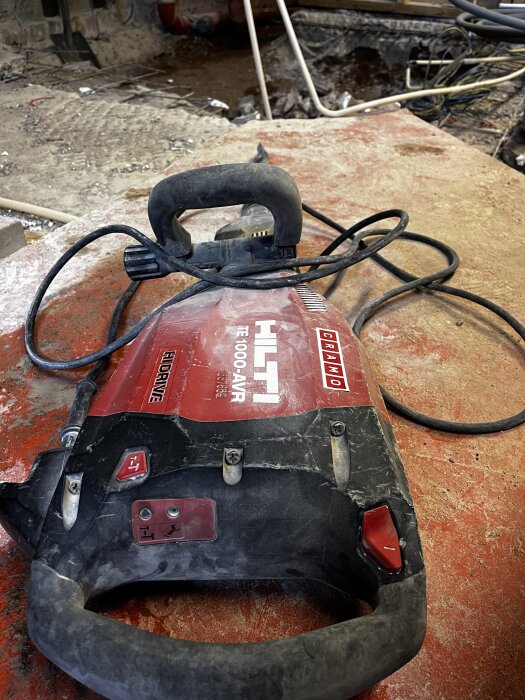 En röd och svart Hilti-kombihammare TE 1000-AVR ligger på ett smutsigt bord med sladd. Bakgrunden visar en byggarbetsplats med rör och betong.